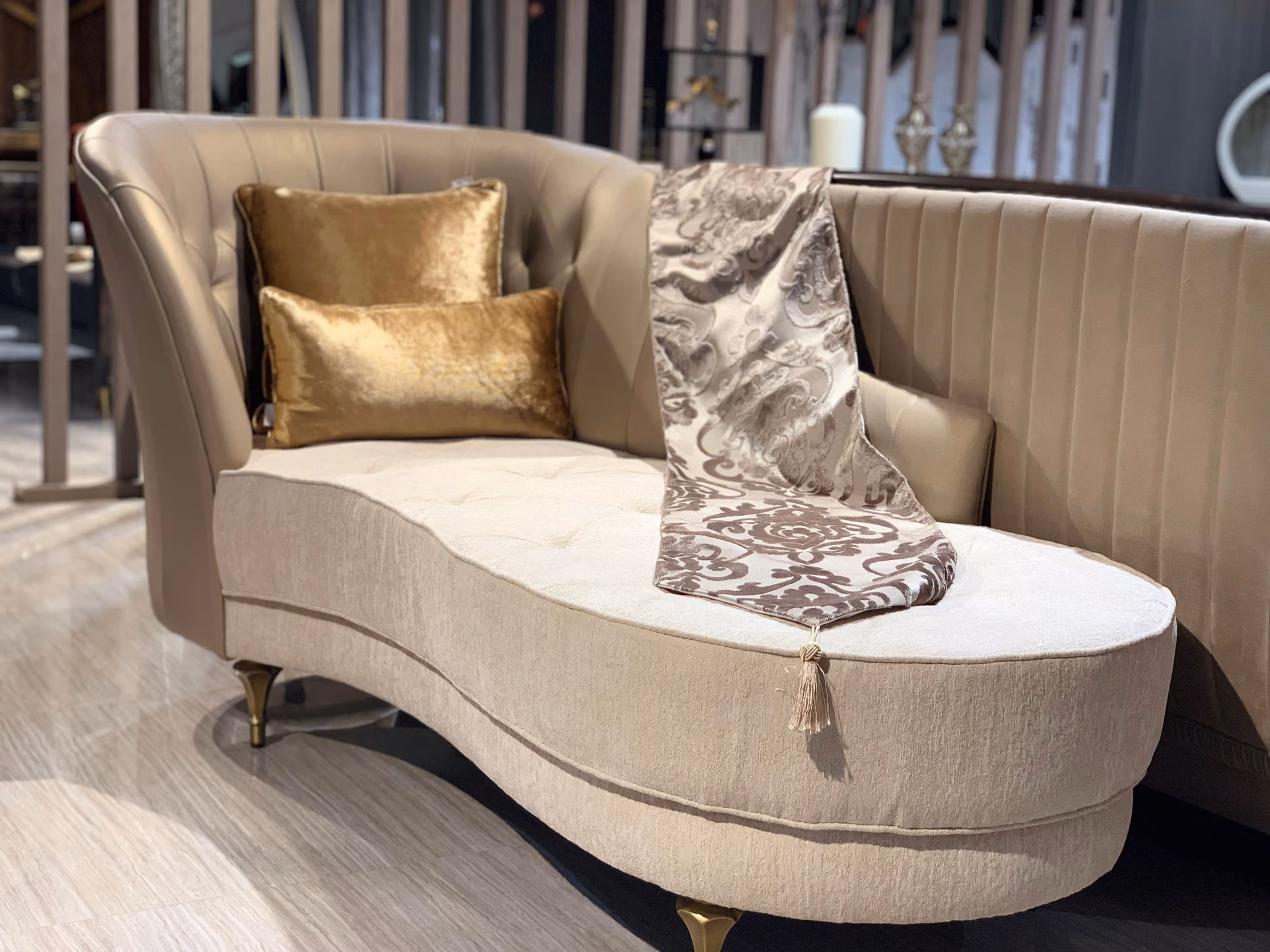 Curved kidney-shaped chaise with a fluted champagne back, cream tufted seat, gold velvet cushions and slim brass legs on a light wood floor.