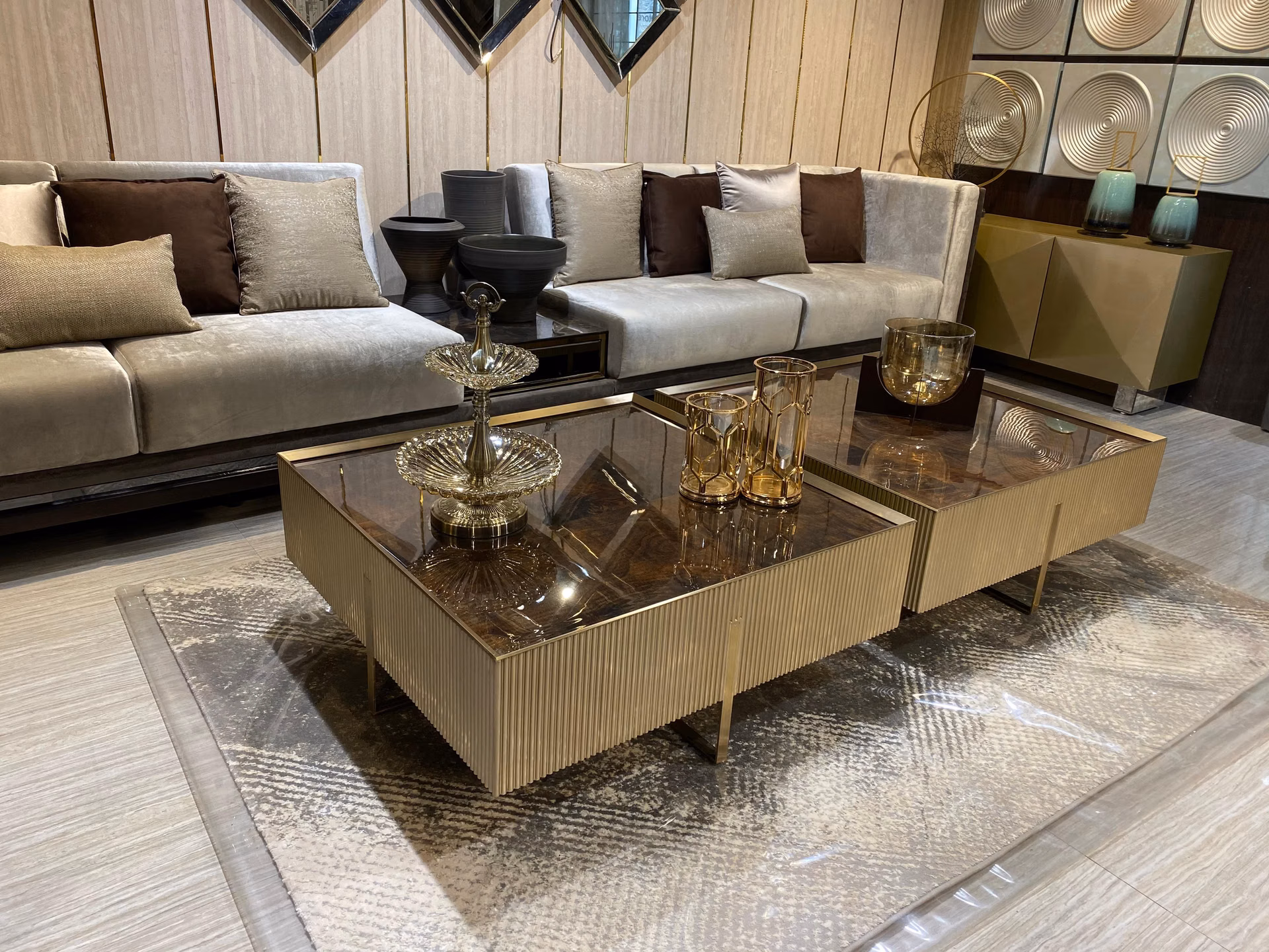 Two-tier round coffee table with a glossy burl-wood and glass top over a fluted champagne-gold pleated drum base, staged in front of a grey sofa.