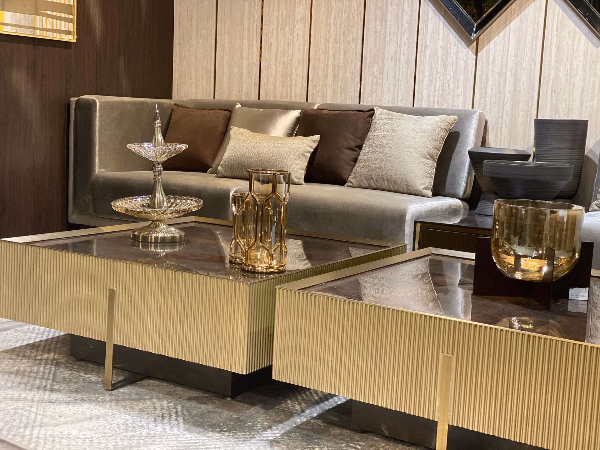 Two square fluted champagne-gold coffee tables with burl-wood and glass tops standing side by side in front of a grey upholstered sofa.