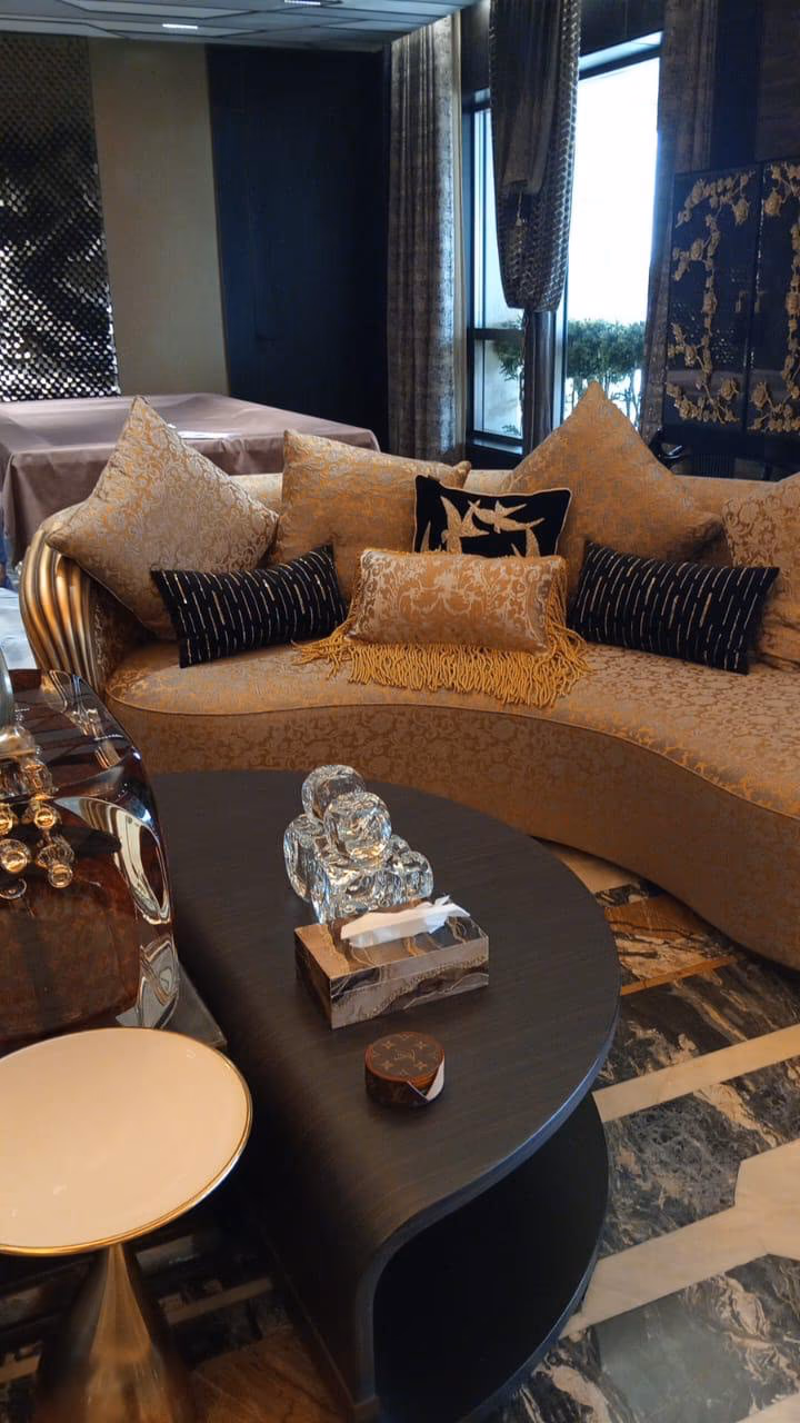 Curved gold damask sofa with a fluted gold-leaf side panel, matching bolster cushions and black beaded pillows beside a dark oval coffee table.
