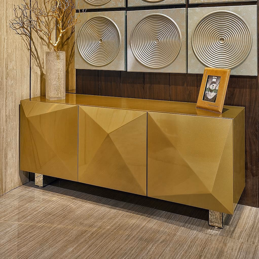 Three-door sideboard in high-gloss gold with a deep faceted diamond-cut front, standing on short antiqued metal legs below a set of concentric-circle wall panels.
