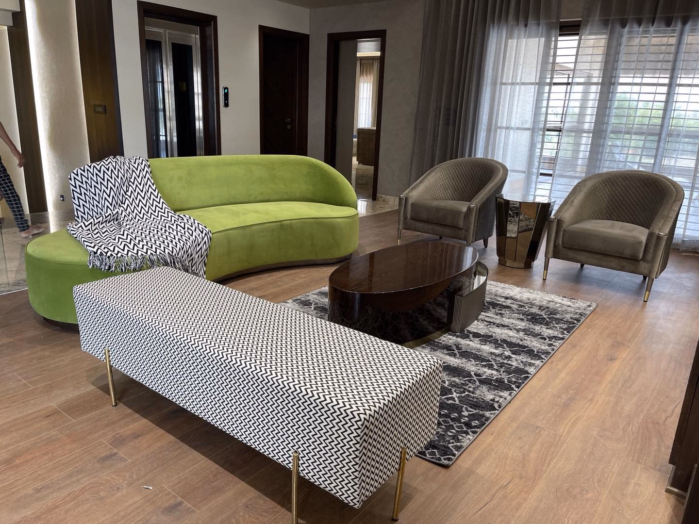 Lounge set with a lime green velvet curved sofa, two taupe channel-back tub chairs on brass rings, an oval high-gloss walnut coffee table and a long chevron-print bench on brass legs.