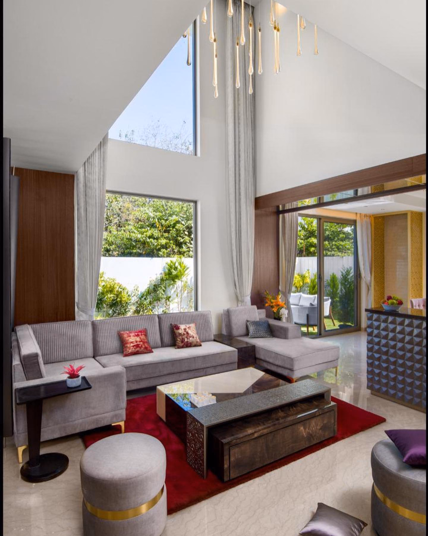 Long grey corduroy L-shaped sectional sofa with a chaise return and brass feet, in a double-height living room with a stone and walnut coffee table, round brass-banded ottomans and a red rug.