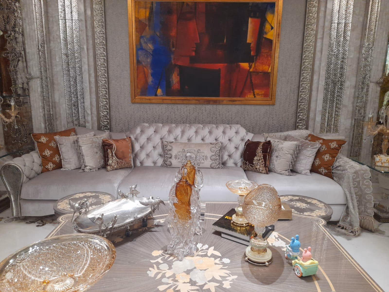 Long low grey velvet sofa with a deep button-tufted back and rows of silver and rust embroidered cushions, framed by silver curtains and wall art.