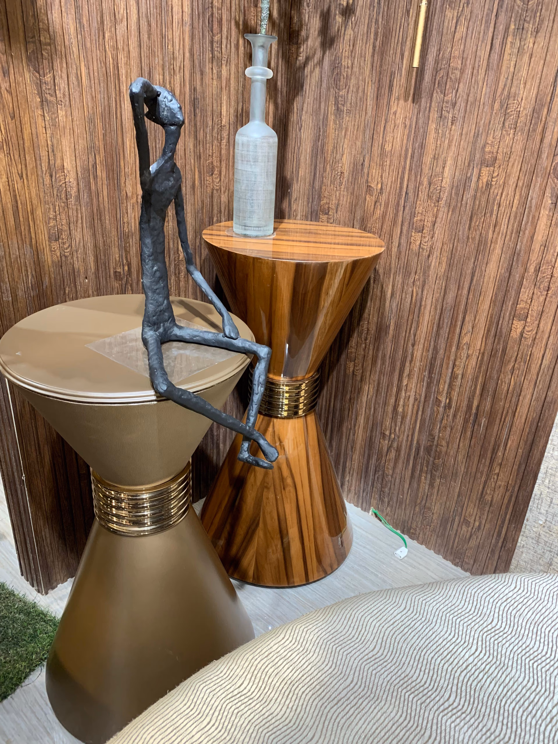 Two hourglass-shaped drum side tables, one in glossy rosewood and one in matte champagne bronze, each split by a ribbed brass band at the waist.