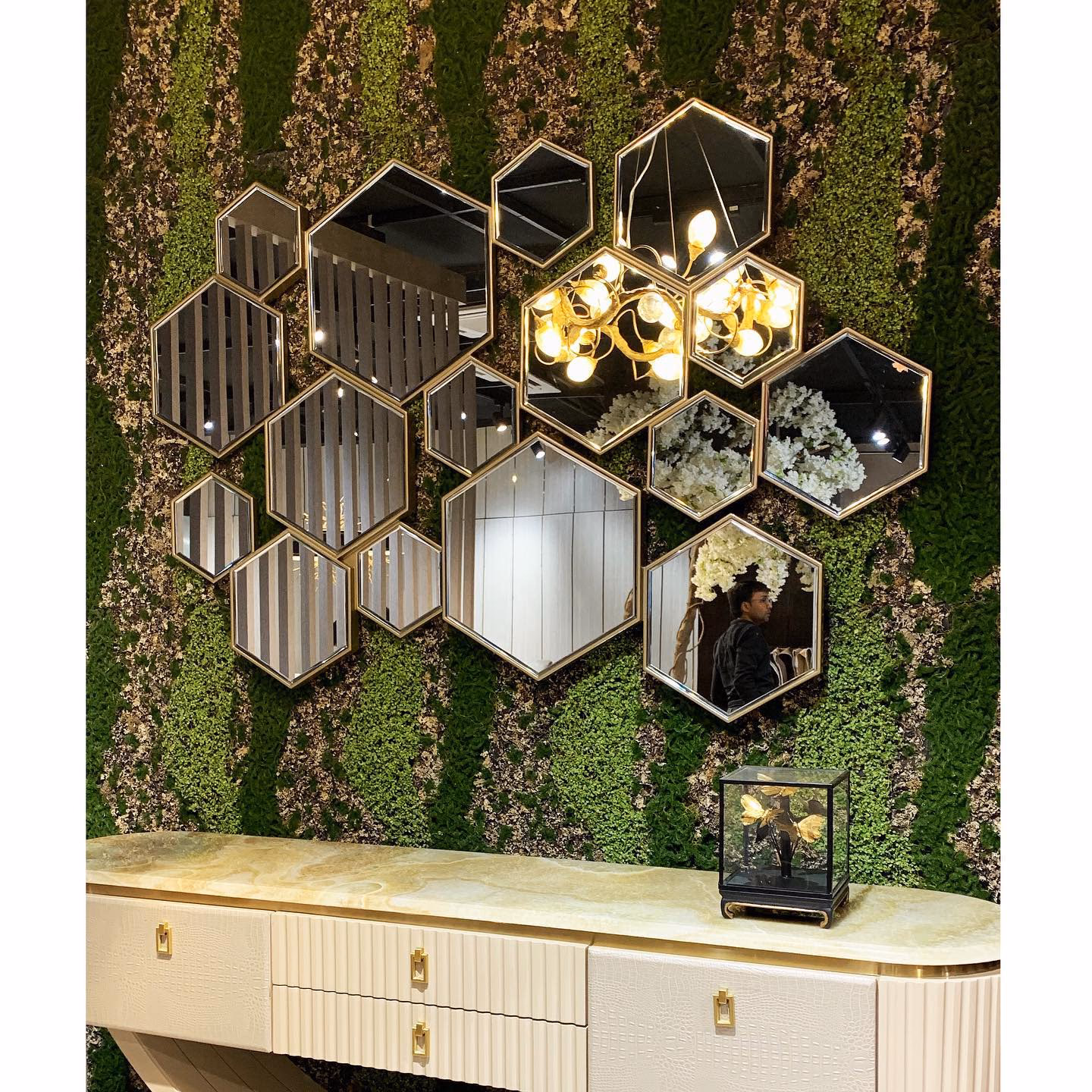 Long console with a backlit green onyx top, ivory crocodile-embossed drawer fronts, fluted end panels and brass drop pulls, set under a cluster of hexagonal wall mirrors on a moss wall.