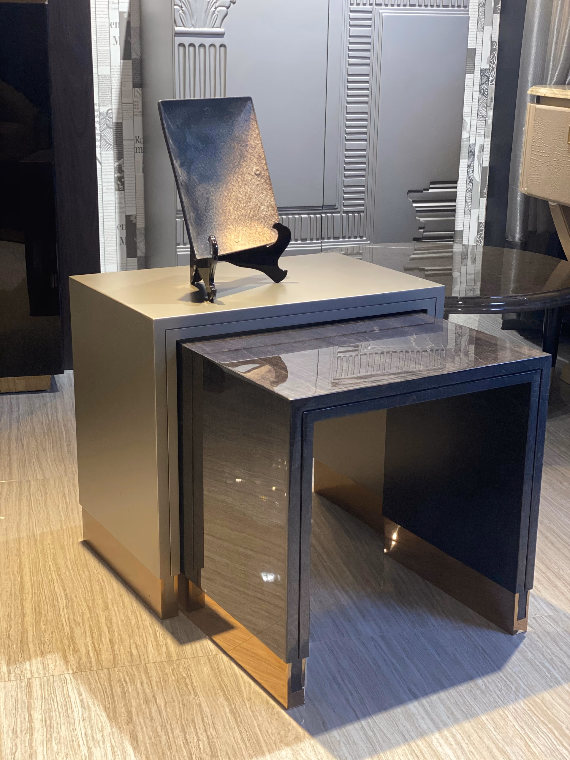 The nesting side tables partly separated, showing the smaller grey stone-grain table sliding out from under the taupe lacquer cube on a wood floor.