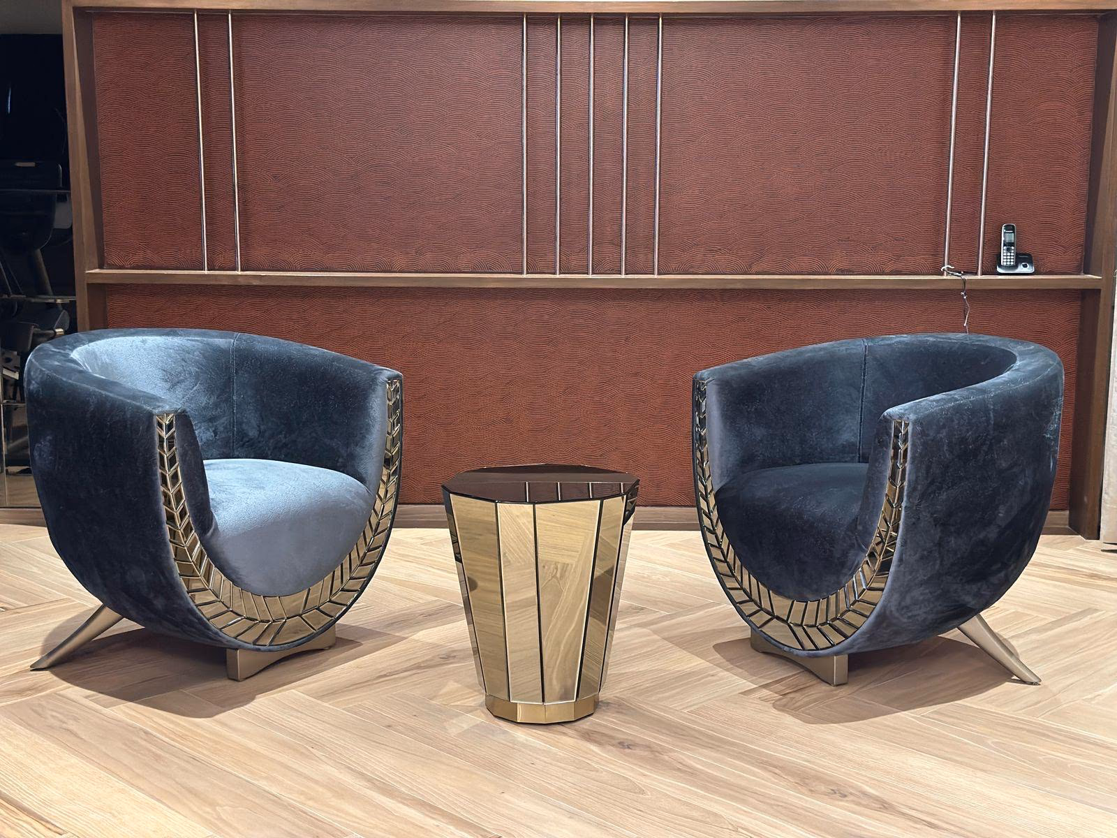 Pair of charcoal velvet barrel lounge chairs with gold leaf-pattern side inlays and angled brass legs beside a faceted gold drum table.