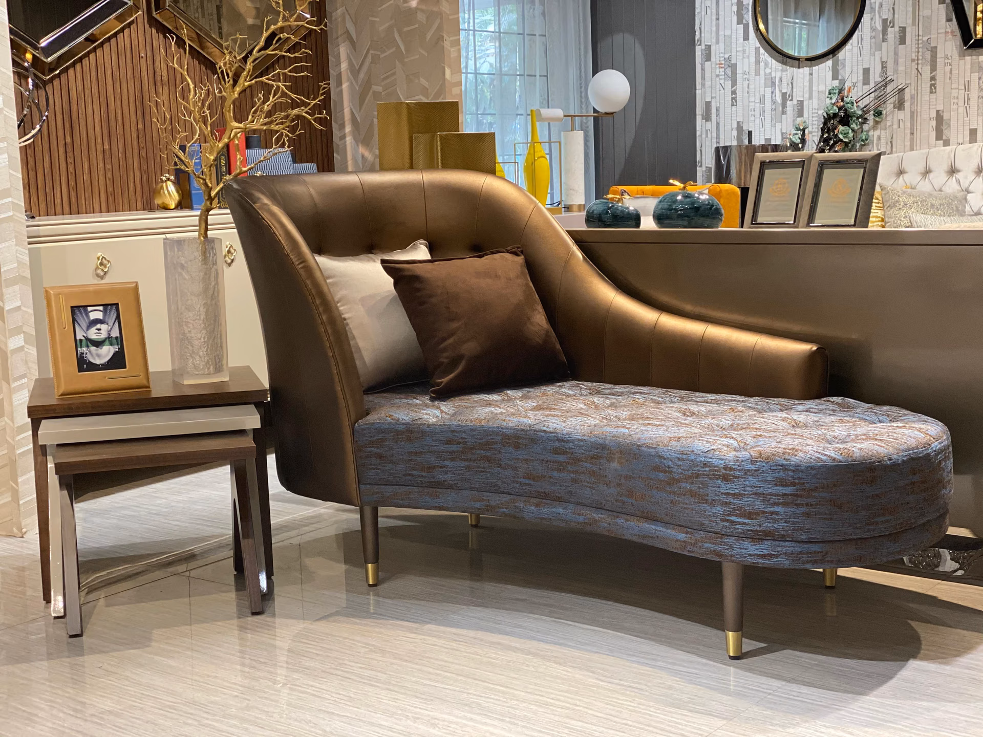 Curved lounge chaise with a smooth bronze-tan leather back curling around a deep button-tufted teal seat, raised on angled dark wood legs.