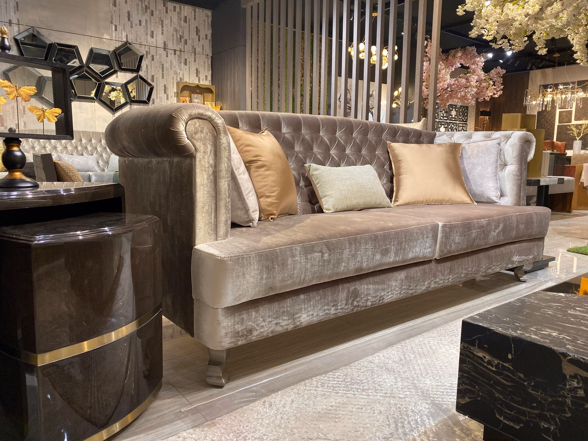 Three-seat chesterfield sofa in soft mink-taupe velvet with deep button tufting on the rolled arms and back, resting on short silver-grey wood feet.