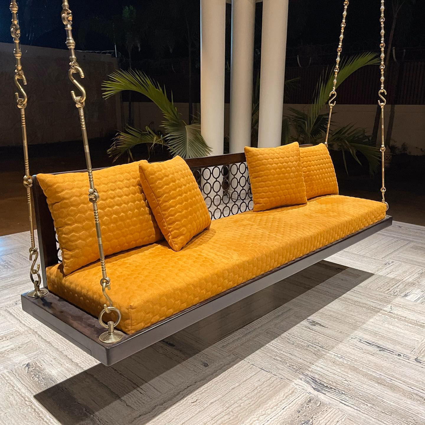 Teak jhula swing seat hung on ornate cast brass chains, cushioned in mustard honeycomb-quilted velvet with matching back cushions, on a terrace at night.