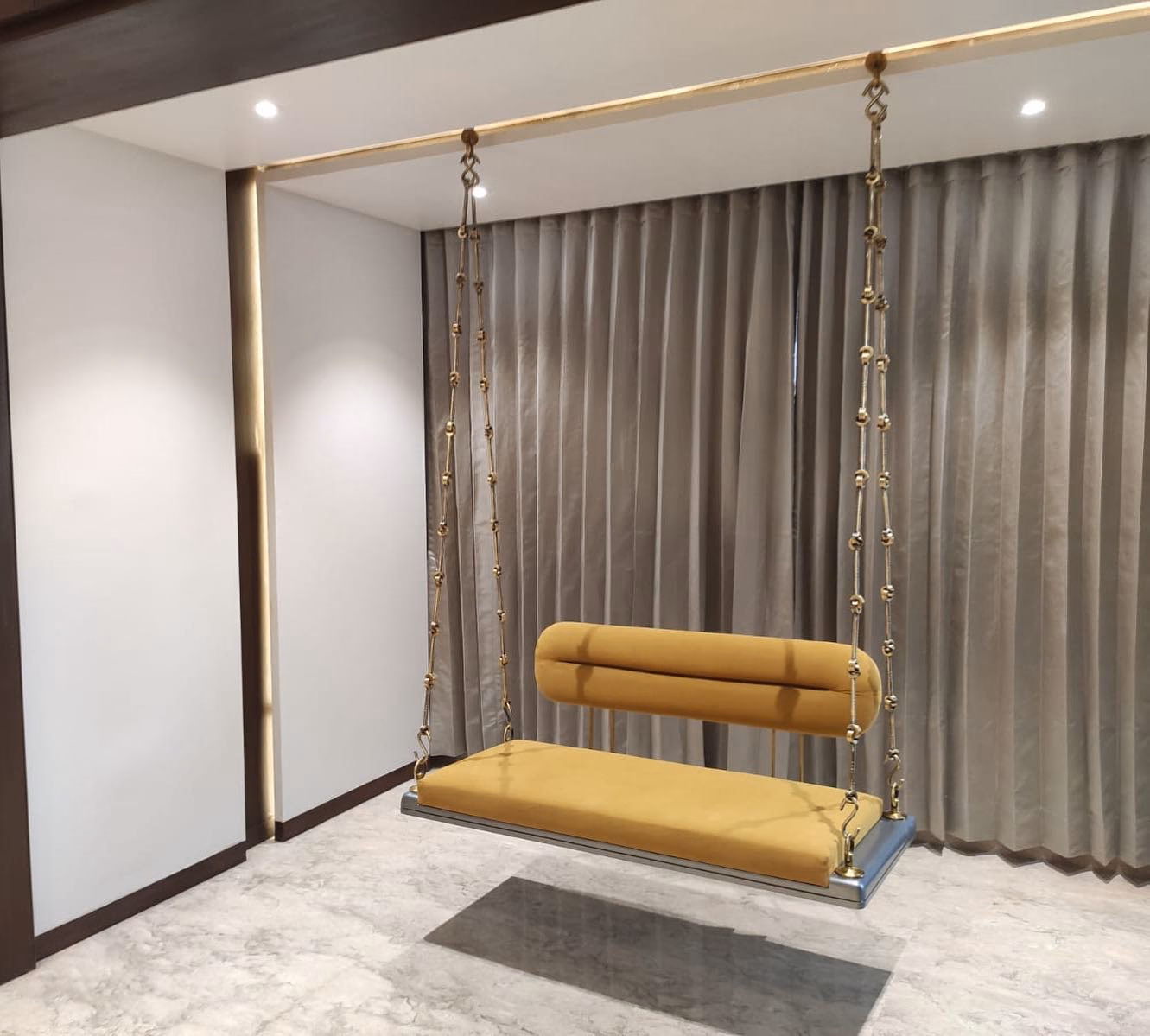 Indoor swing with a mustard velvet seat and a long rolled bolster back, hung from ceiling-fixed decorated brass chains on a steel top rail against grey curtains.