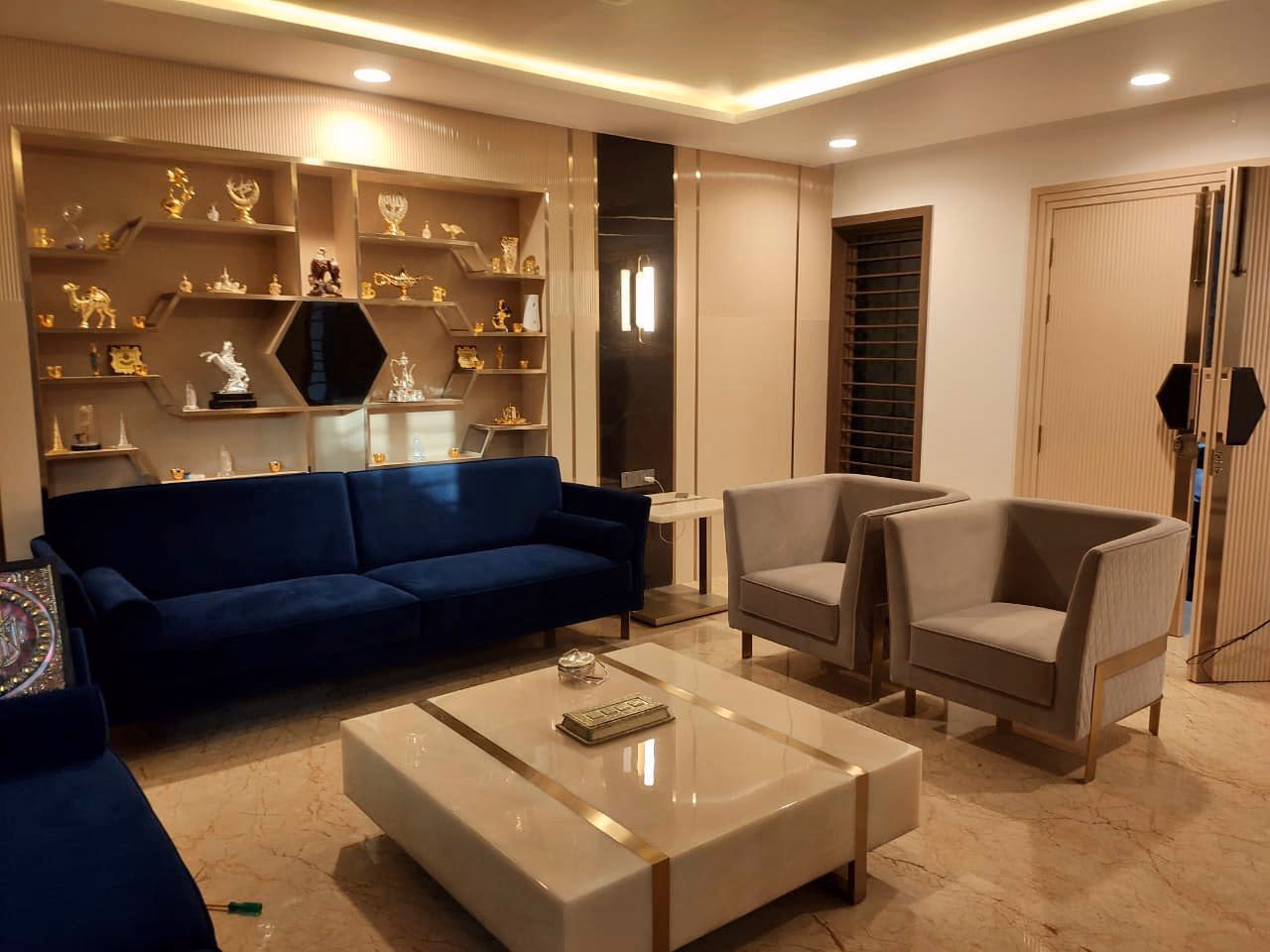 Living room set with a navy velvet three-seat sofa on brass legs, two grey armchairs with gold frames, a low white marble coffee table banded in gold, and hexagonal display shelving behind.