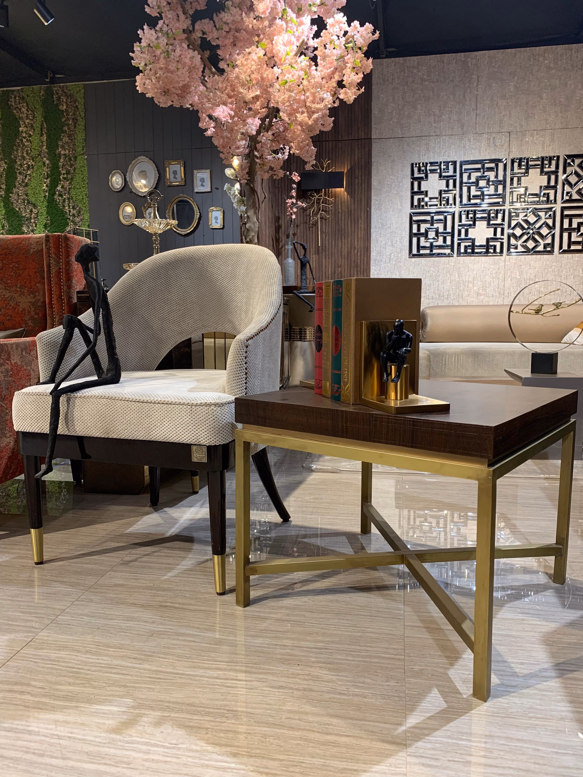 Angled view of the oatmeal woven barrel accent chair beside a dark walnut side table with a slim brass X-frame, under artificial cherry blossom.