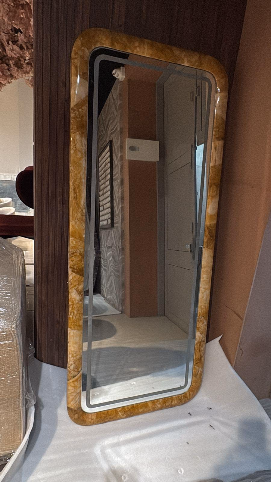 Tall full-length mirror with a wide amber onyx frame and softly rounded corners, leaning against a dark ribbed wood wall.