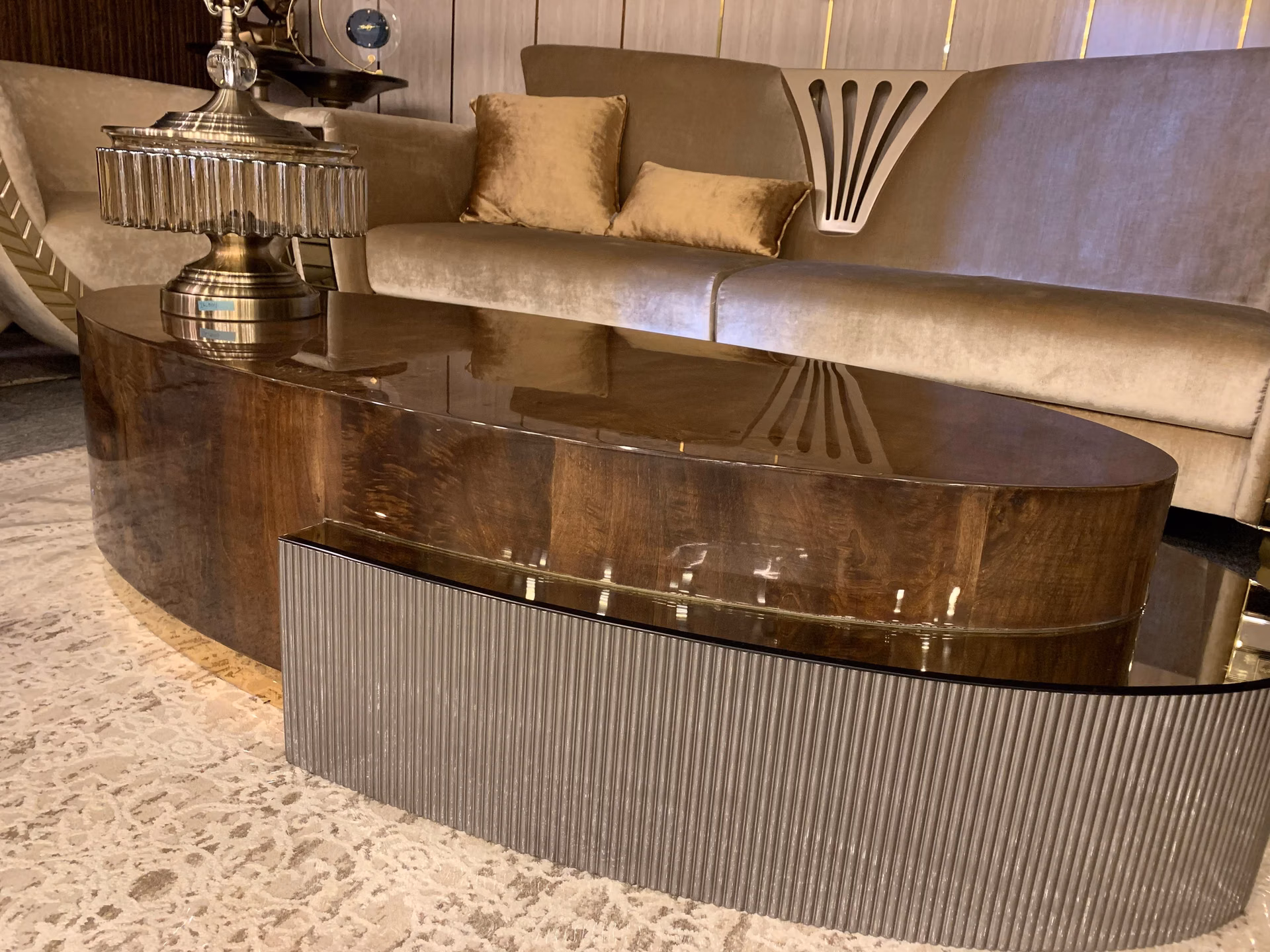 Angled view of the oval burl coffee table showing the deep fluted pewter base curving around its full length, with a ribbed glass lamp on the polished top.