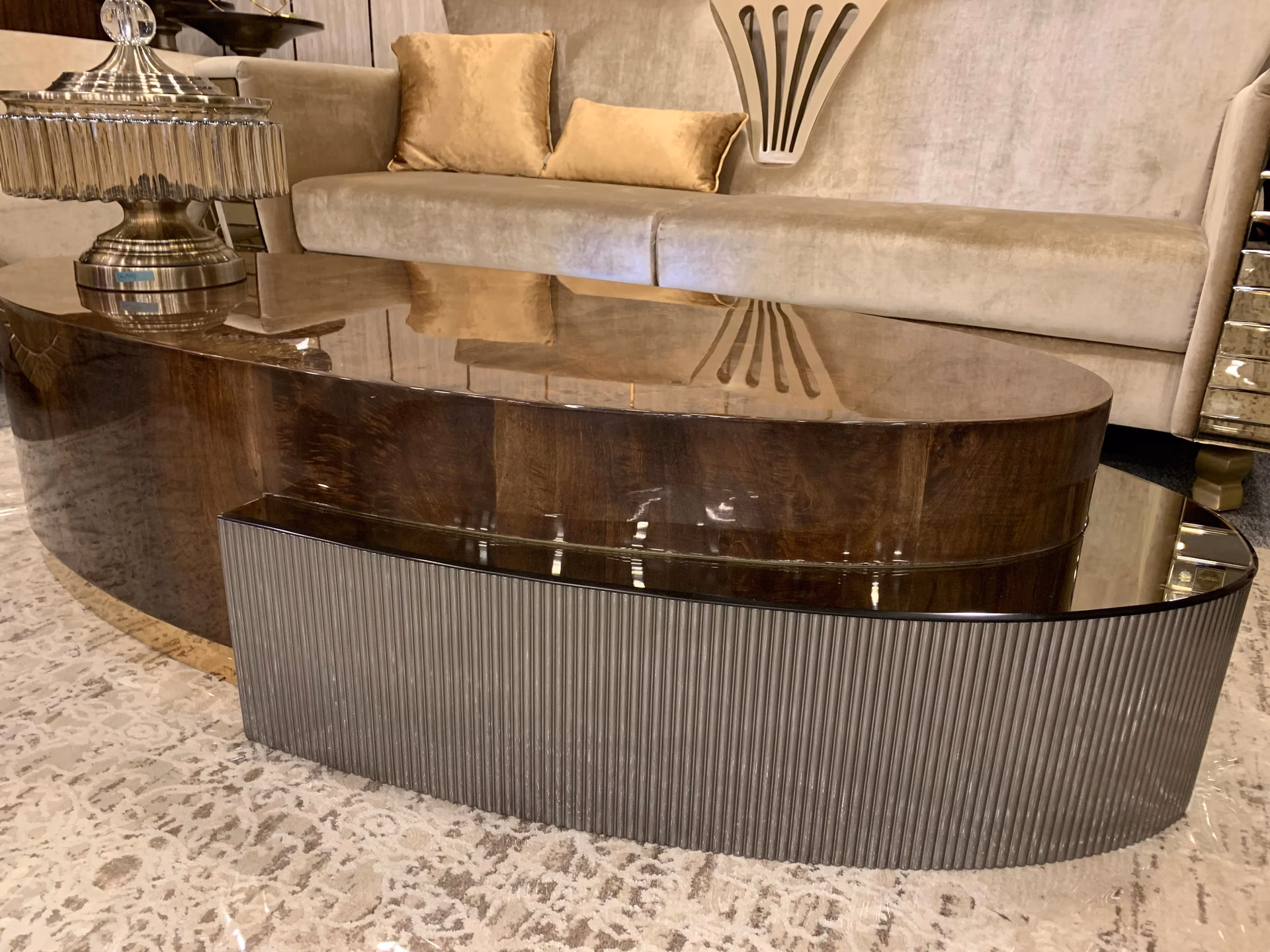 Long oval coffee table with a high-gloss dark burl top and a fluted pewter-grey base, sitting on a patterned rug before a beige velvet sofa.