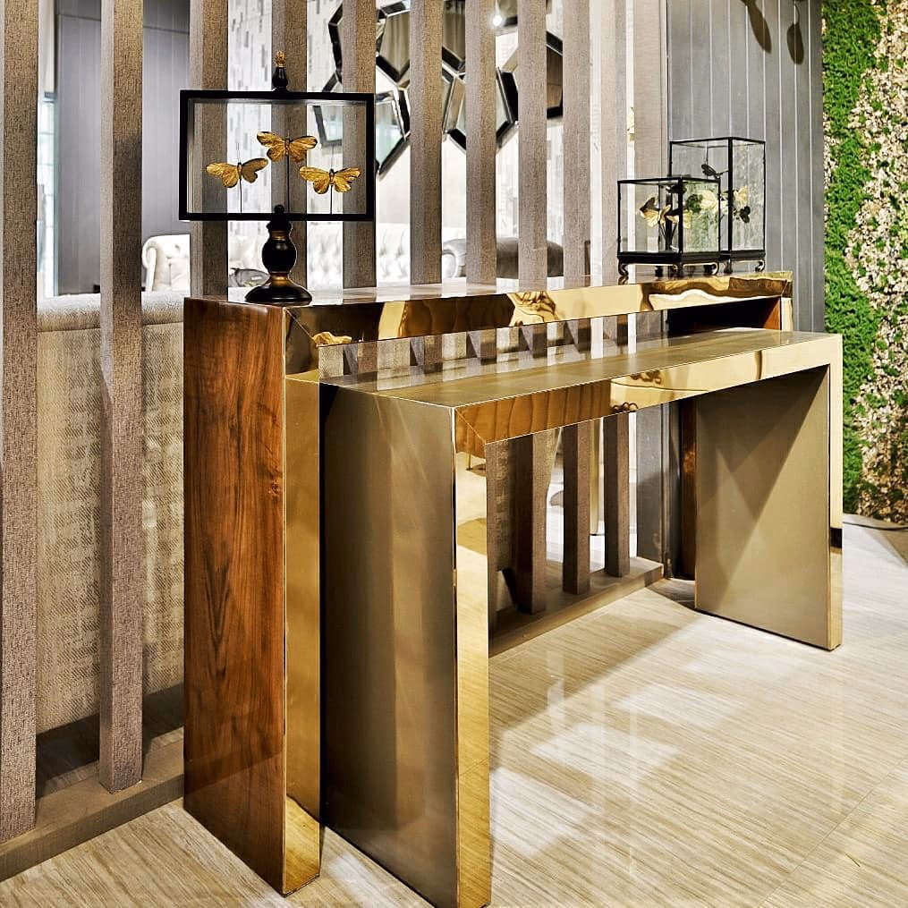 Angled view of the polished brass waterfall console beside its walnut twin, styled with a butterfly dome and a black lamp against a slatted screen and a moss wall.