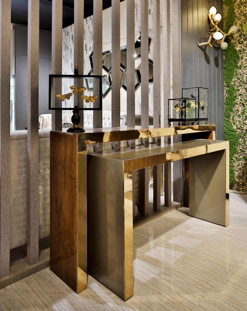 Two nested waterfall console tables in high-polish mirror brass, set against a wood-slat room divider with butterfly display domes on top.