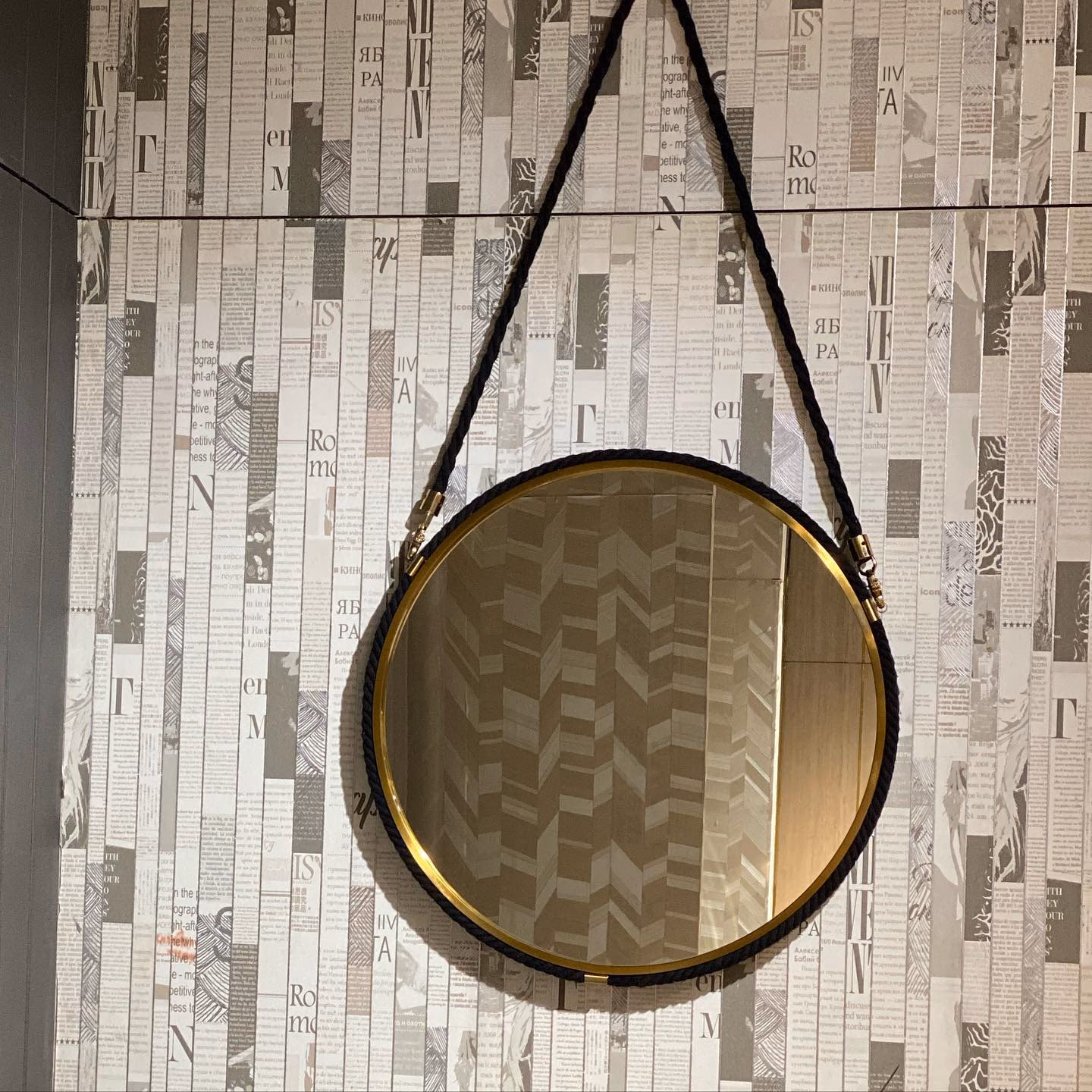 Round wall mirror in a slim polished brass frame, hung from a thick braided black rope fixed to two brass cleats, against a printed textured wall.