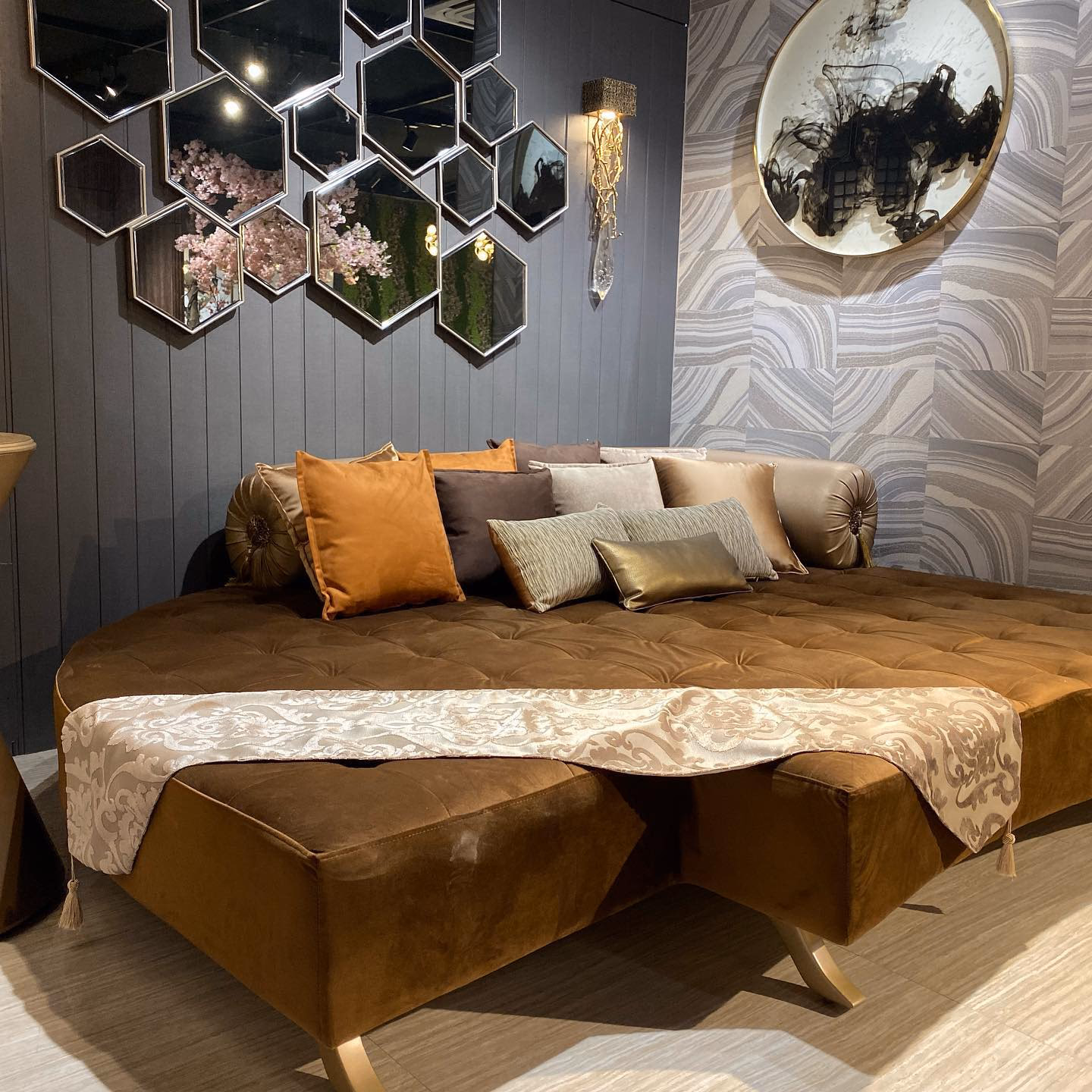 Large round daybed upholstered in tan suede with a deep button-tufted top, a scalloped skirt and splayed brass legs, dressed with bolsters and cushions under a hexagonal mirror cluster.