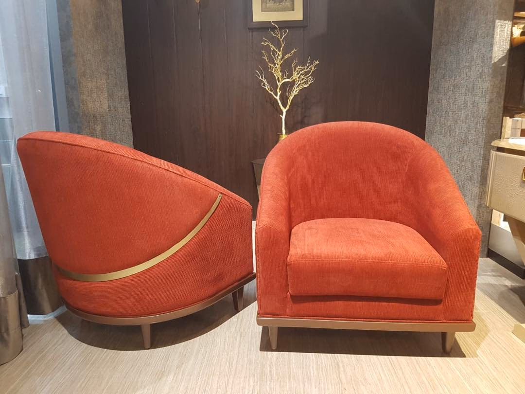 Pair of rust orange corduroy tub chairs, one shown from behind with a curved brass band sweeping across its back, both on low walnut plinth bases.
