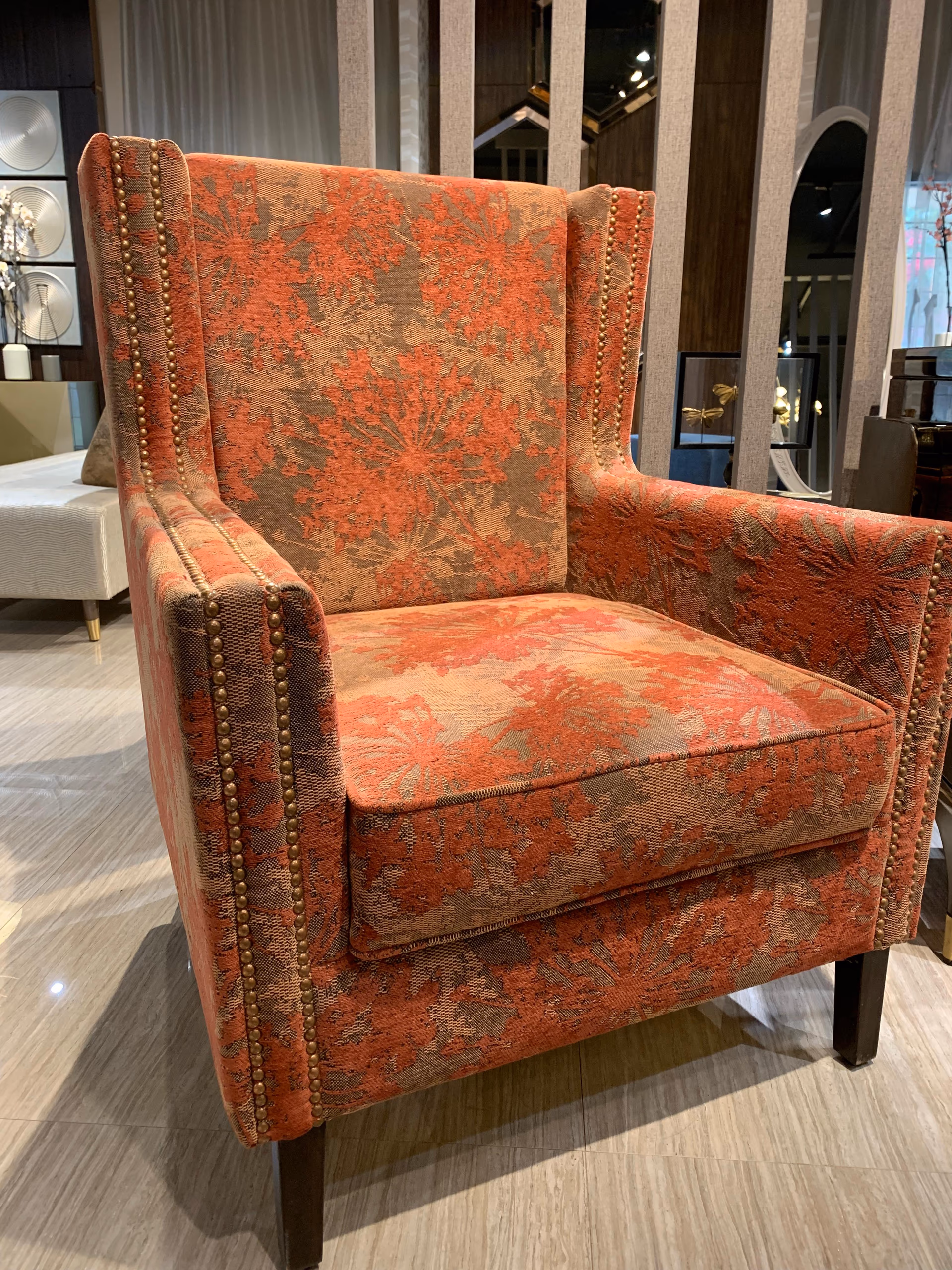 High wingback armchair covered in rust and beige floral chenille with brass nailhead trim along the arms and wings, on dark wood legs in a showroom.