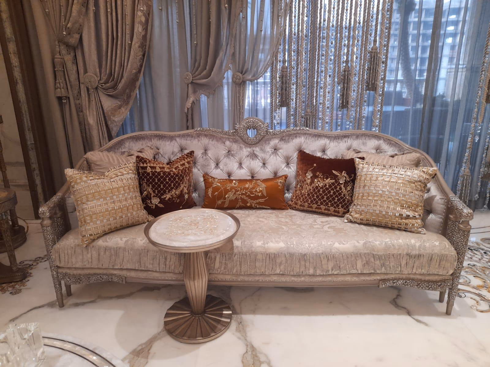 Grey velvet French-style sofa with a camel-back top, central carved silver medallion crest and layered velvet cushions against sheer tasselled curtains.