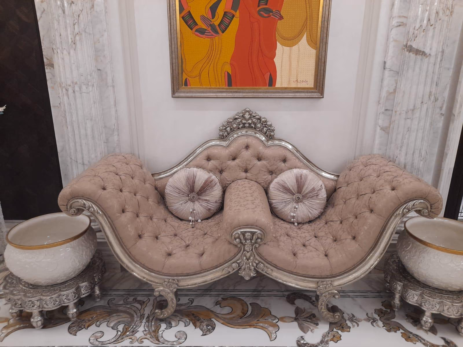 Symmetrical double-scroll settee in blush tufted velvet with a silver-carved wave frame and a rose crest, flanked by two silver-footed urns under bright wall art.