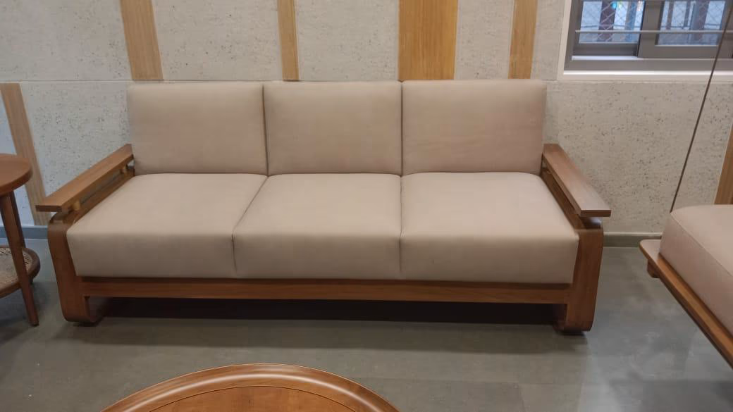 Three-seat sofa with an exposed solid-teak frame, wide flat armrests and thick beige fabric cushions, against a stone-and-wood panelled showroom wall.
