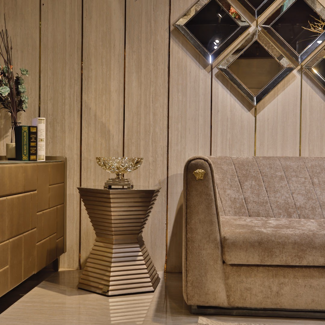 Hexagonal side table built from stacked stepped bands in warm bronze that narrow to a waist and flare out again to the floor, standing beside a channel-tufted sofa.