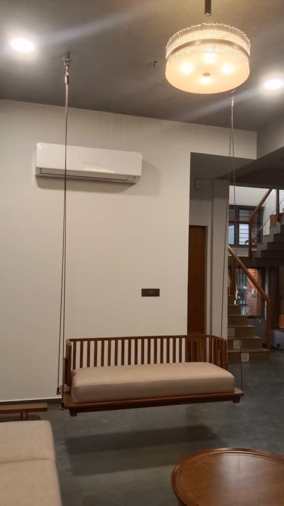 Solid-teak slatted-back swing bench with a long cream cushion, hung from the ceiling on thin steel cables near a staircase and a crystal drum chandelier.