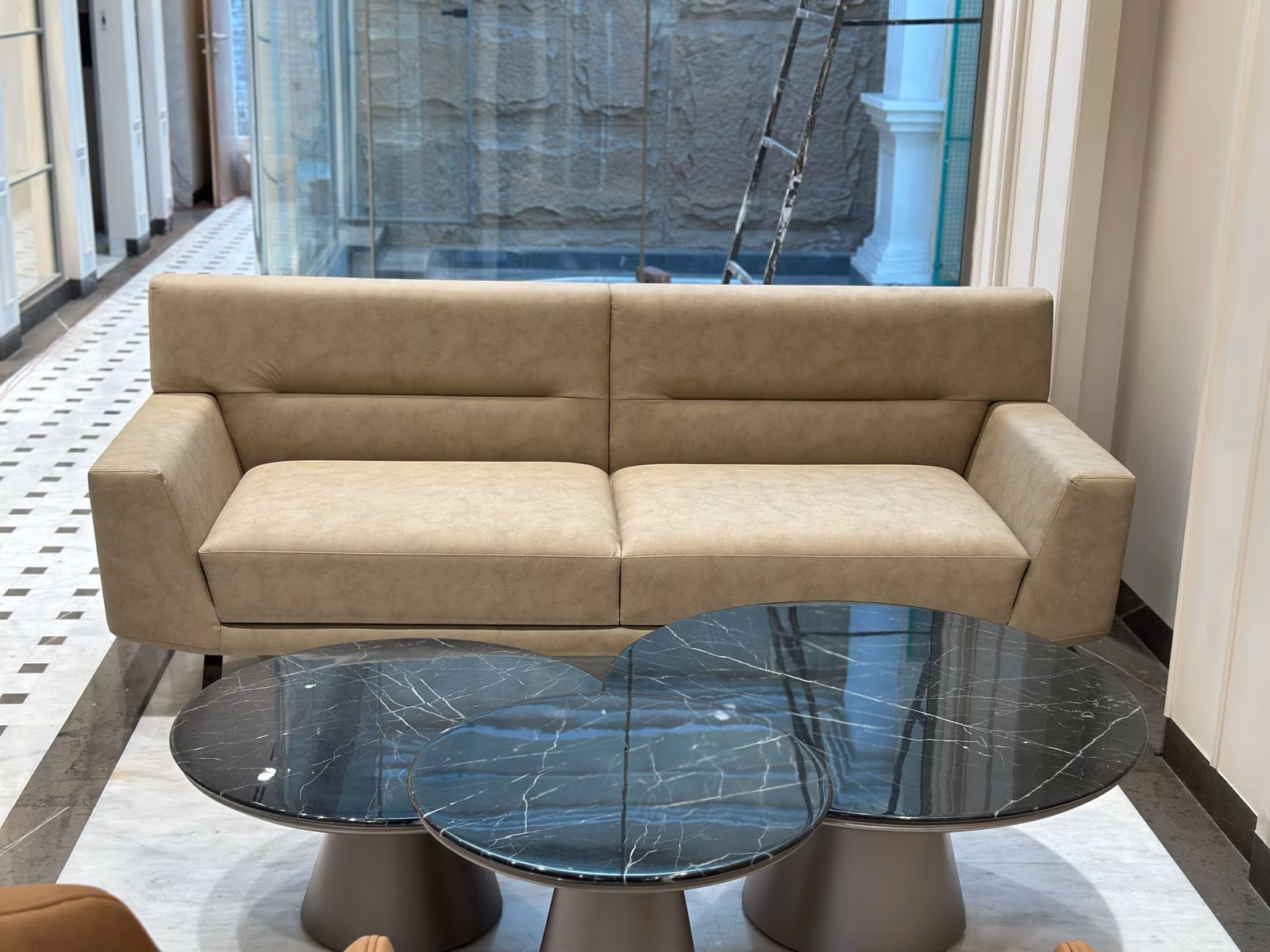 Taupe leather two-seat sofa with squared block arms and tufted low back beside a trio of black marble nesting tables.