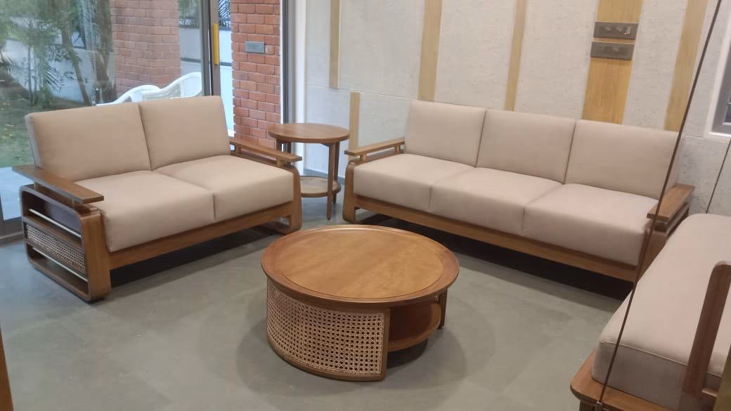 The teak sofa collection arranged as a set, a two-seater and three-seater in beige with a round cane-wrapped teak coffee table between them by a brick wall.