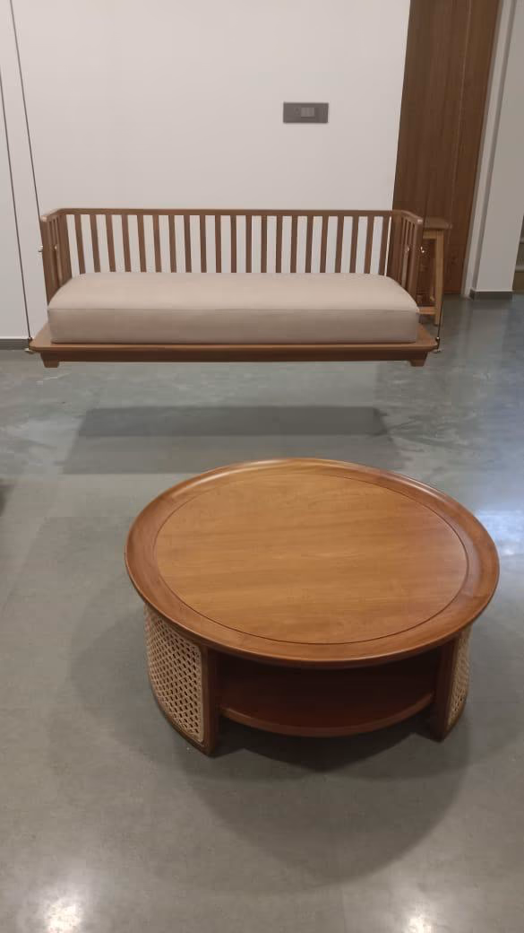 The slatted teak swing bench resting against a wall with its cream cushion, staged in front of the round cane-and-teak coffee table on a concrete floor.