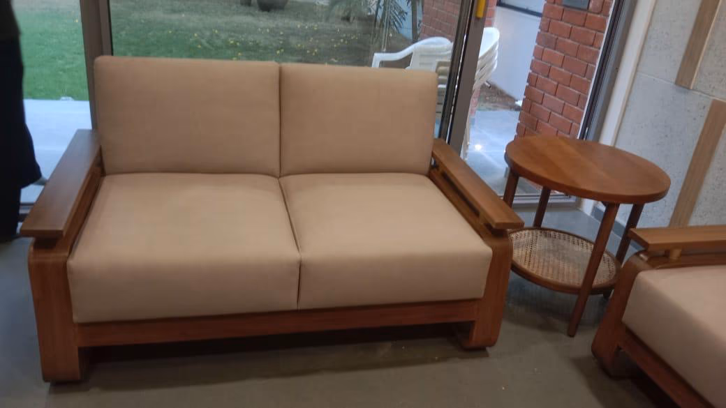The solid-teak sofa in a two-seat size with wide flat arms and beige cushions, beside a small round cane-shelf side table by a garden window.
