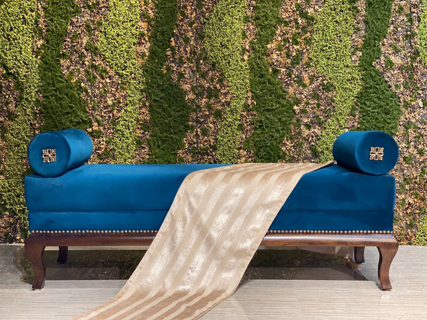 Long teal velvet bench with a bolster cushion at each end held by carved gold rosettes, antique brass nailhead trim along the rail and carved dark wood cabriole legs.