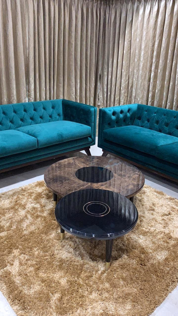 Two matching teal-green velvet sofas with deep button-tufted backs facing a round two-tier coffee table on a patterned rug in a styled living room.
