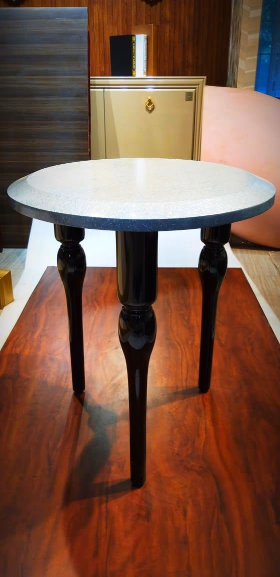 Small round side table with a speckled cream-and-grey terrazzo top on three glossy black turned legs, standing on a polished wood floor.