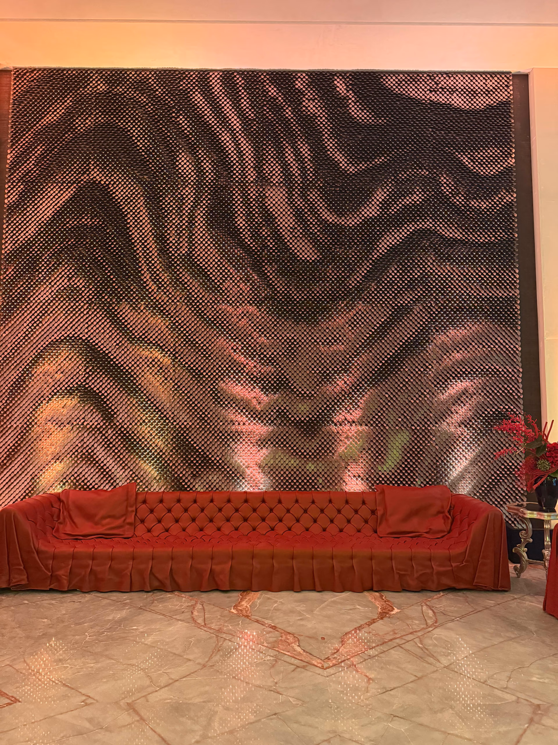 Long red tufted sofa with a pleated skirt and two bolster cushions set against a large scale-pattern mosaic feature wall.