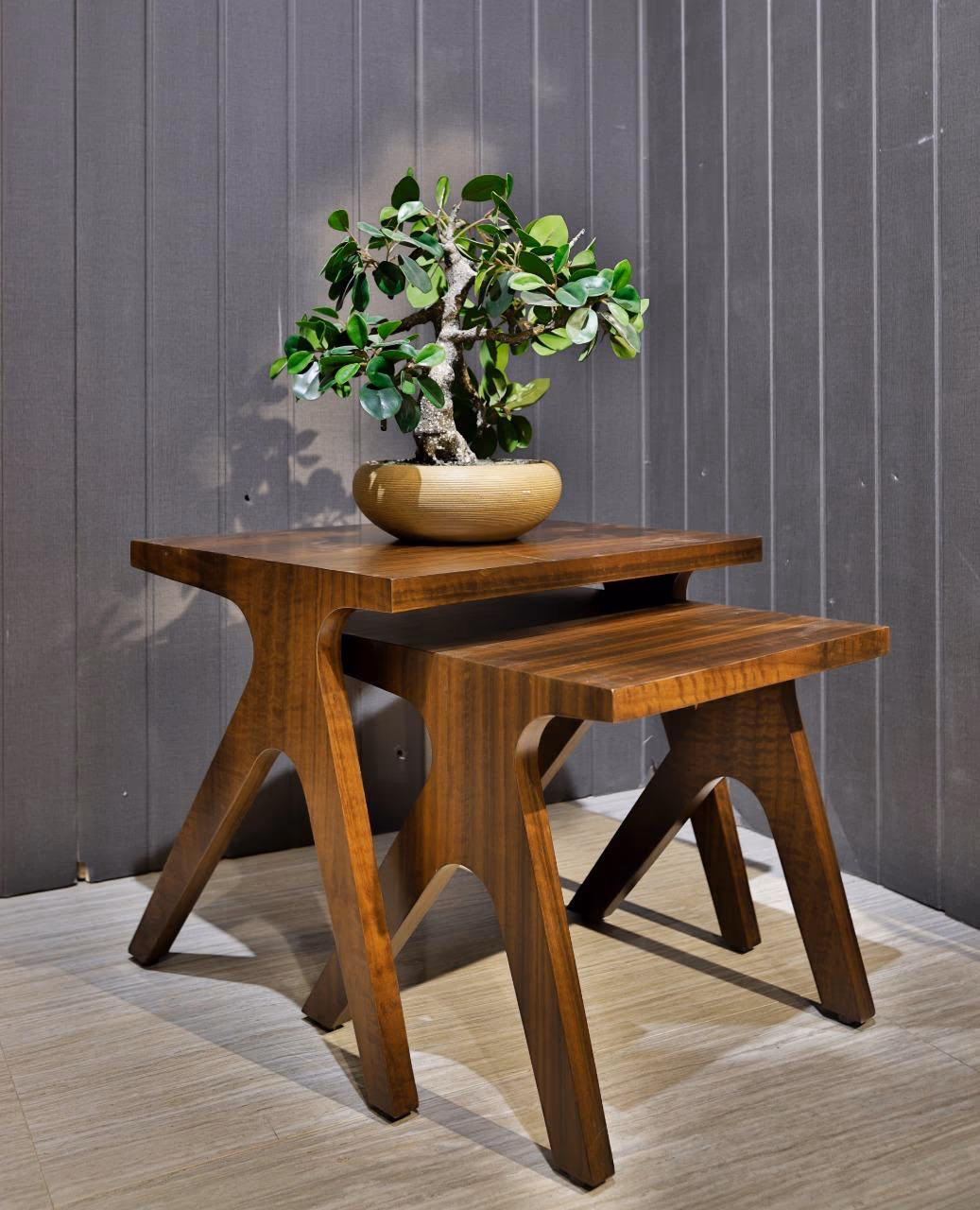 Pair of solid walnut nesting side tables with clean tops and angular splayed A-frame legs, holding a bonsai against a grey panelled wall.