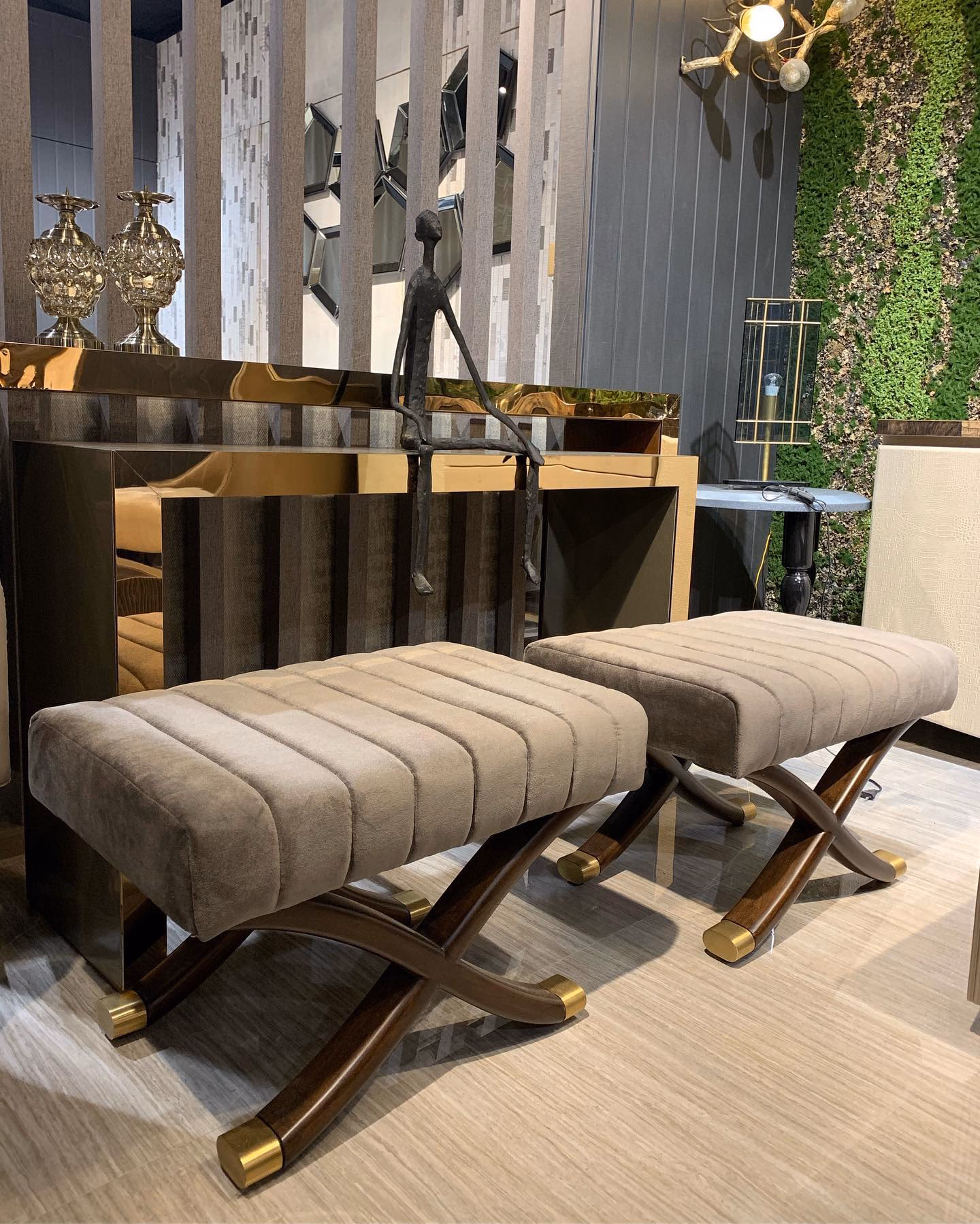 Pair of benches with taupe velvet channel-tufted seats on curved walnut X-frames finished with polished brass foot caps, standing in front of a bronze mirrored console.
