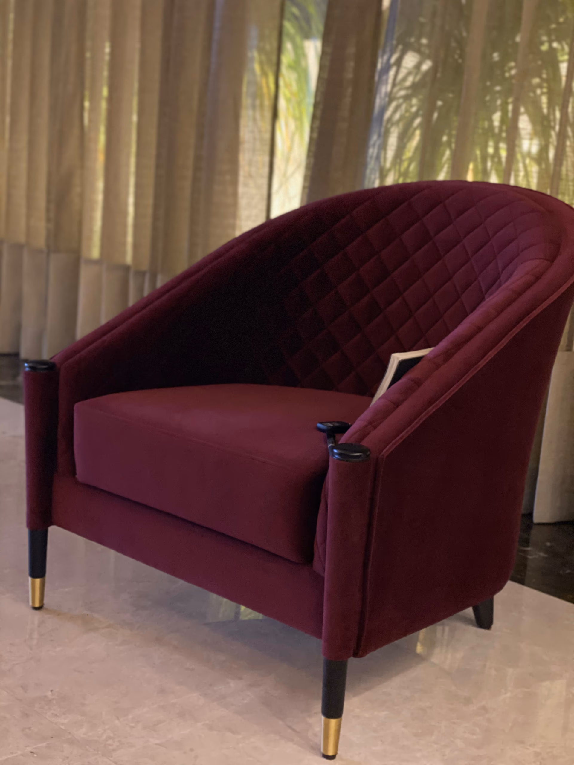 Deep wine velvet tub chair with a curved diamond-quilted back, plain seat cushion, black lacquered arm caps and black tapered legs with brass tips, before sheer curtains.