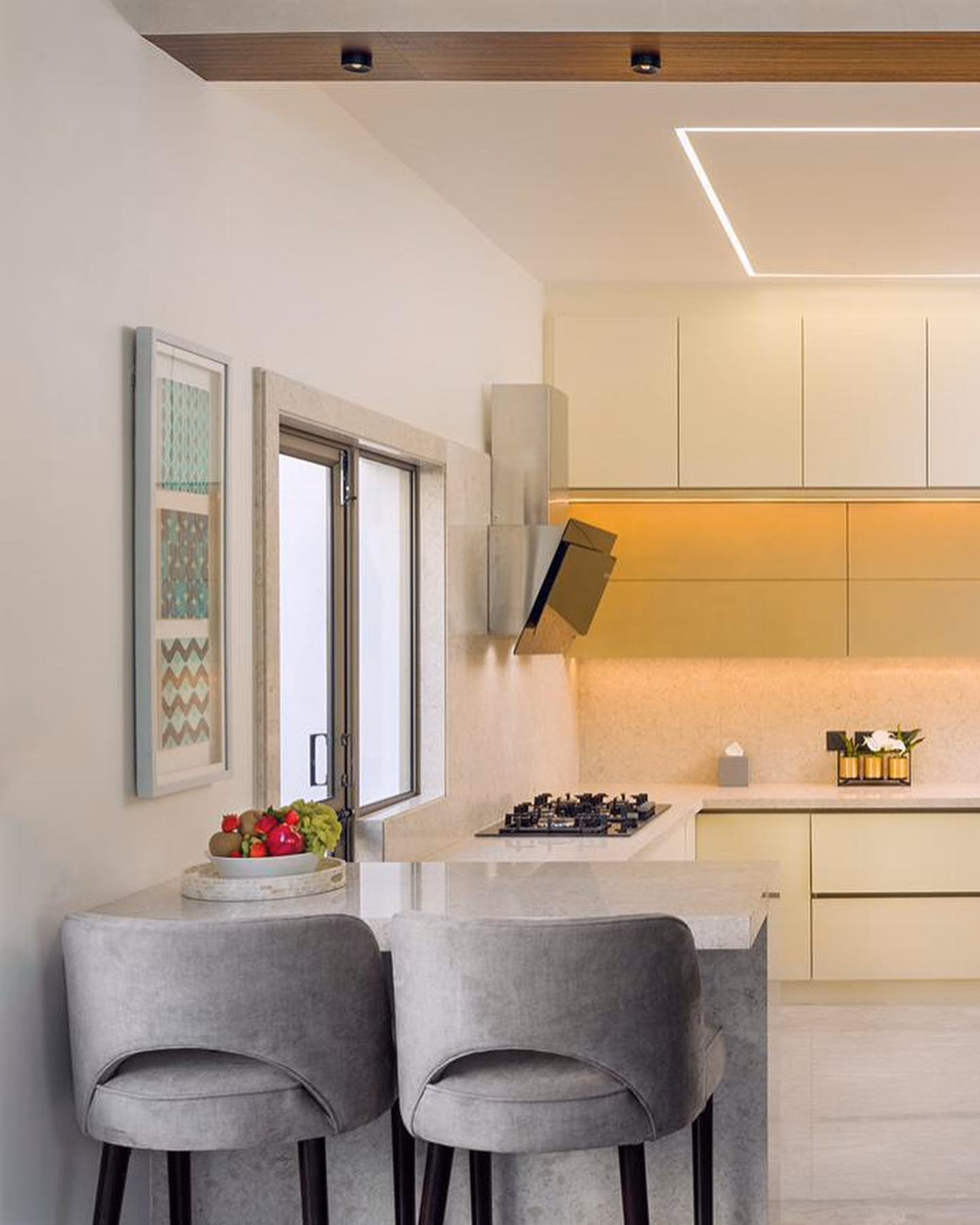 Compact cream handleless modular kitchen with an angled black extractor, under-cabinet lighting and a pale marble breakfast counter with two grey velvet bar stools.