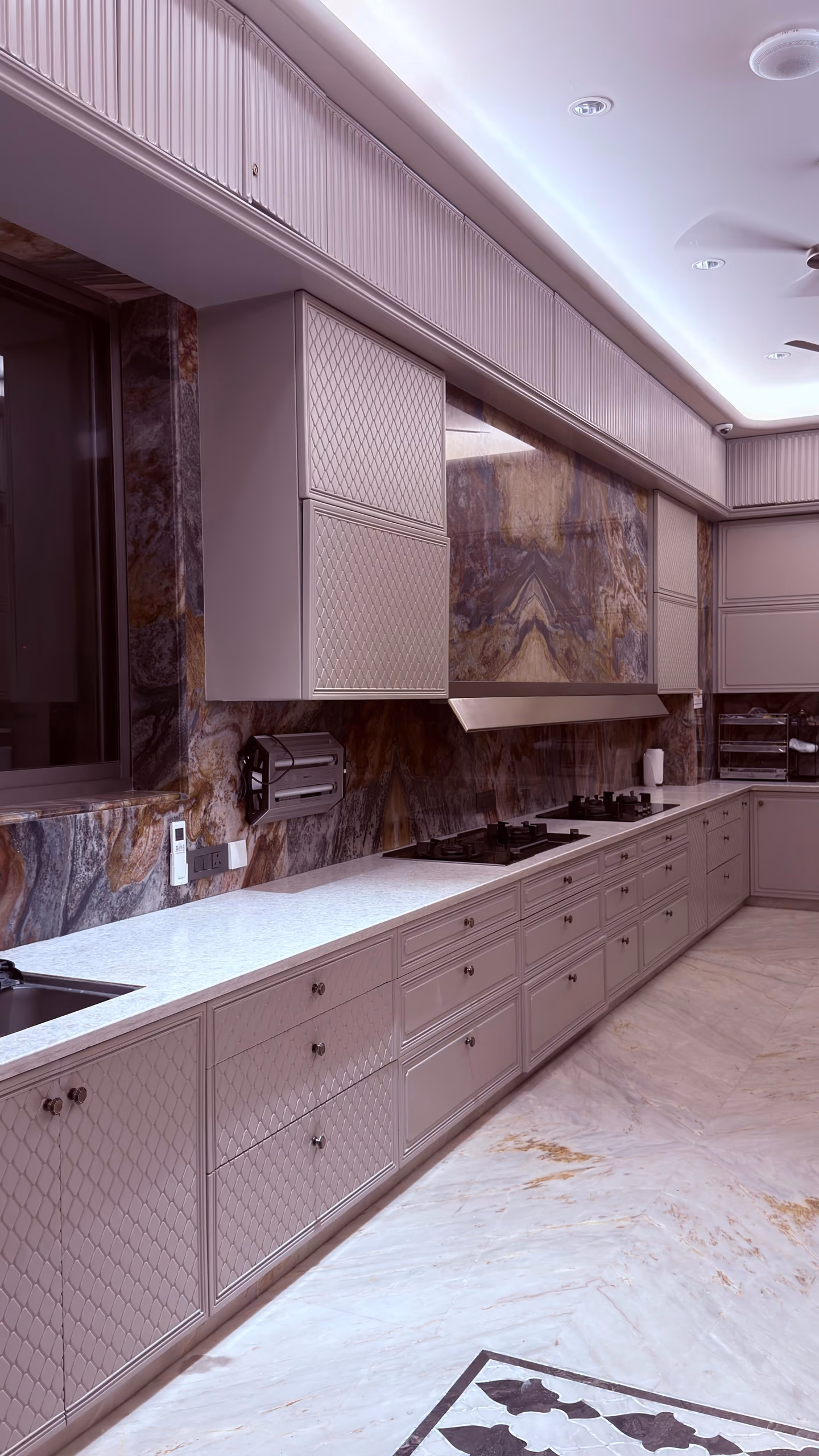 L-shaped modular kitchen with taupe fluted and diamond-textured cabinets, brass knobs, a dramatic brown-and-blue marble backsplash, hob and slim chimney.