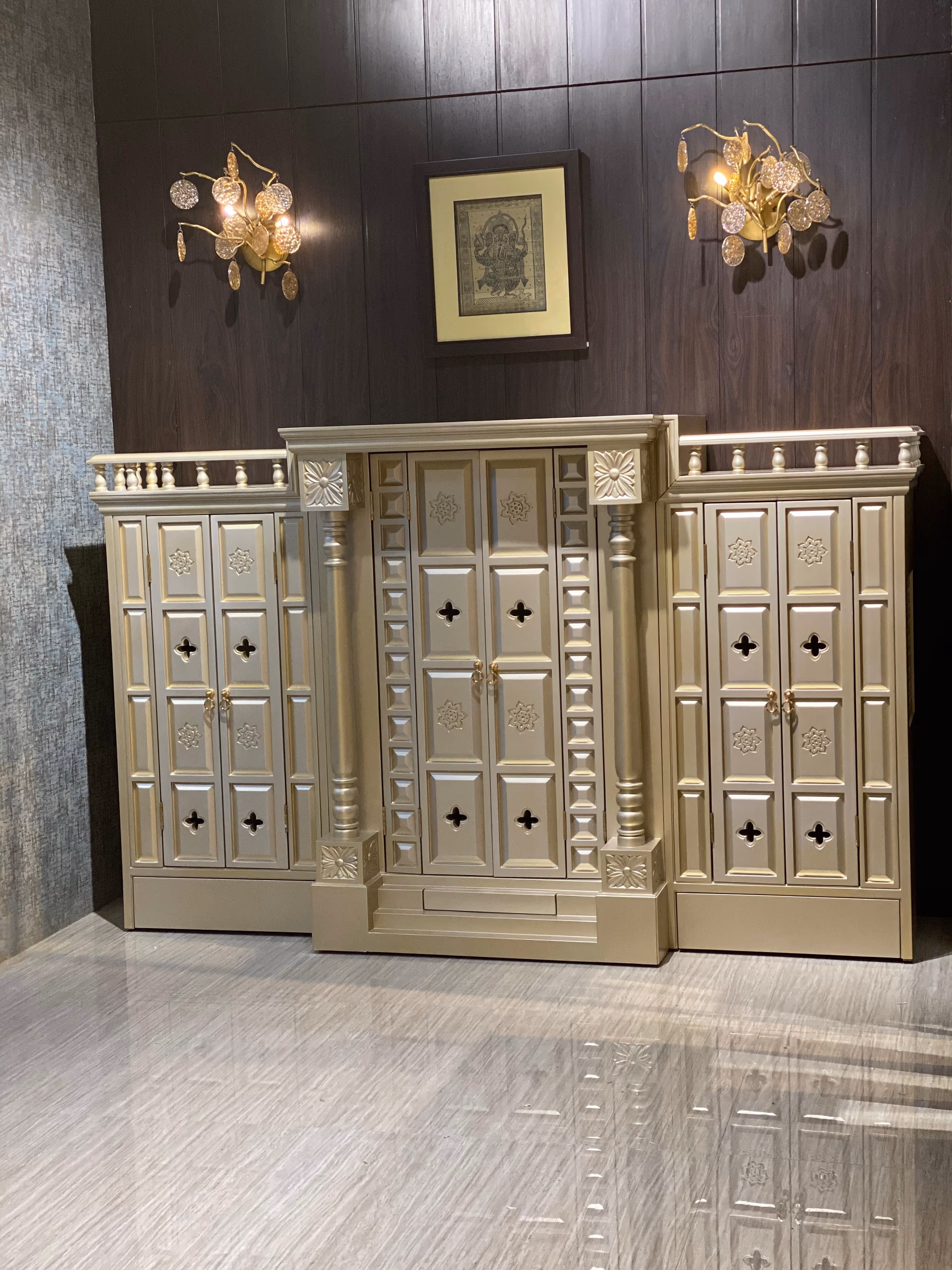 Champagne-gold wooden temple with two carved pillars, cusped doors bearing lotus rosettes and pierced quatrefoil vents, flanked by matching cabinets under a Ganesha print.