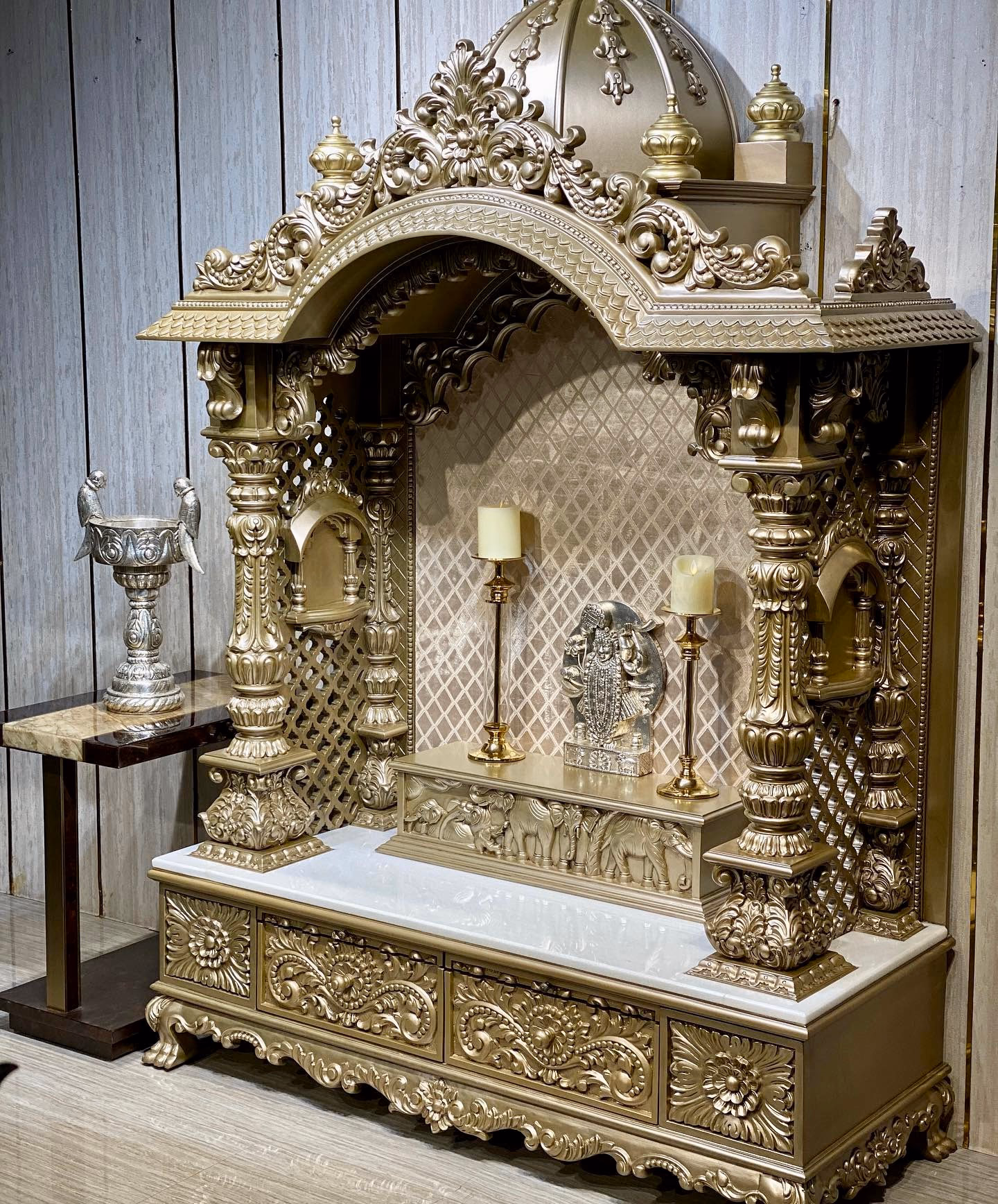 Champagne gold pooja mandir with a carved dome and kalash finials, deep scrolled arch, pierced jaali side panels, carved elephant frieze and a white marble platform over a carved drawer chest.