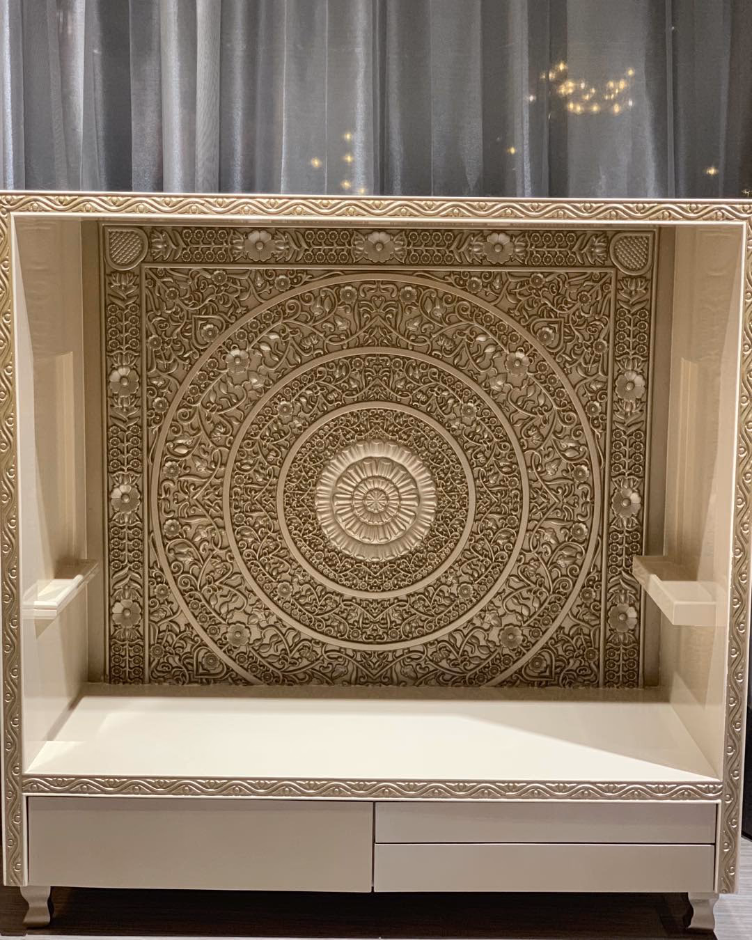 Ivory pooja mandir with a deeply hand-carved concentric mandala back panel of lotus and floral bands, a beaded carved surround, a clear white platform and two storage drawers below.