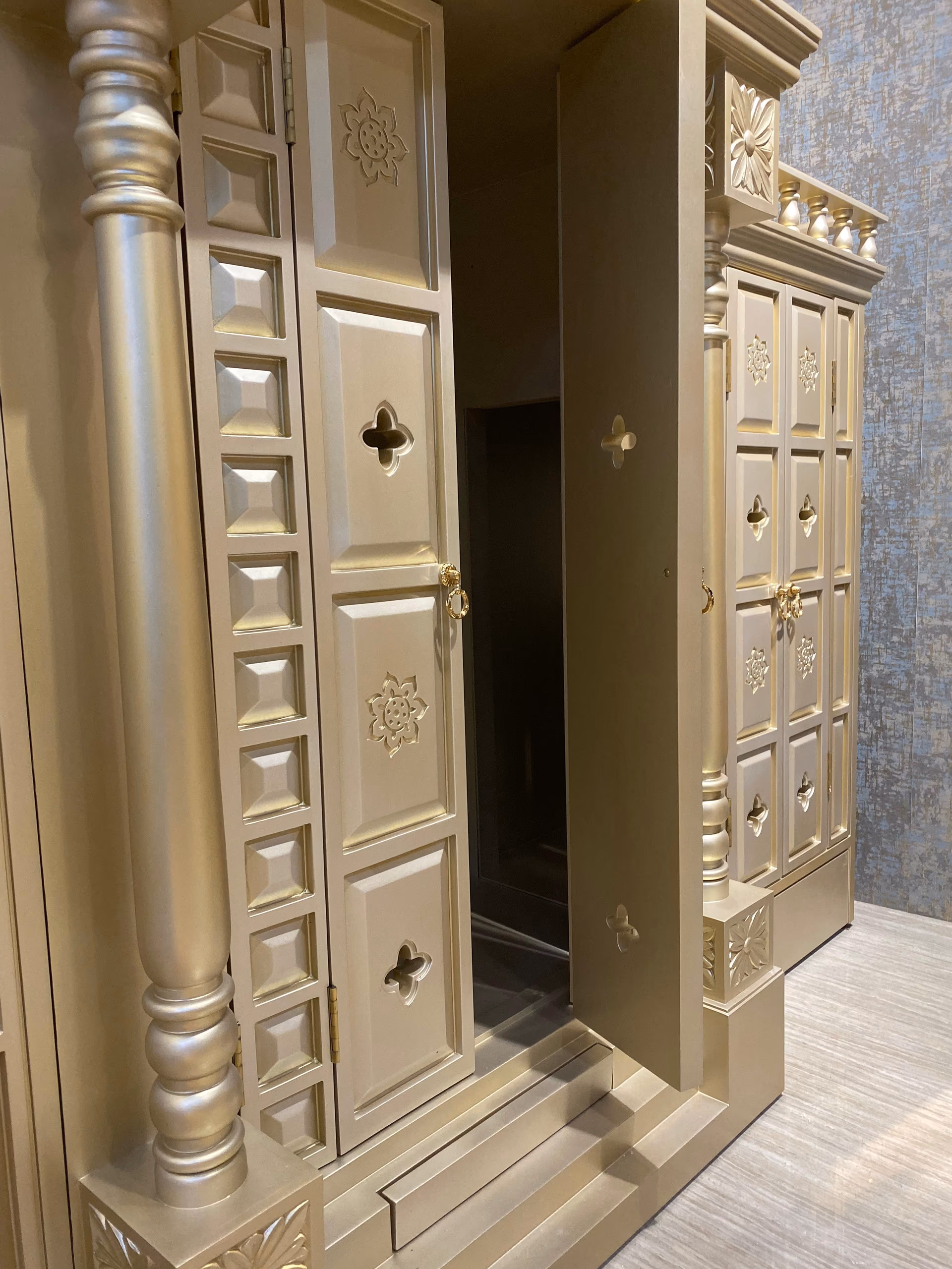Angled view of the mandir with one carved door open, showing the beaded pillar, pierced quatrefoil cut-outs and the shrine space inside.