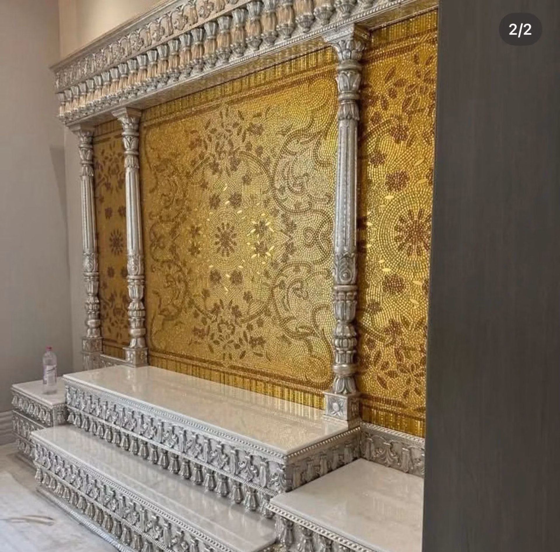 Grand silver hand-carved pooja mandir with fluted silver pillars, a stepped white-marble platform and a floral gold-mosaic back wall in warm amber tones.